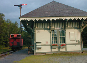 Highly decorative one-story frame building painted in cream and olive green set on a gravel lot next to a portion of train track upon which a red train car sits. A train signal is attached to the building.