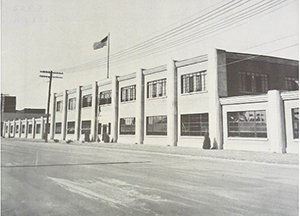Black and white image of stepped concrete building with a two-story core flanked by one-story wings. Building is lit by large multi-pane steel windows.