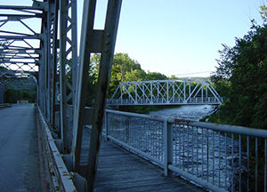 Distant view of a metal truss bridge over a river, surrounded by trees and blue sky. Close-up view of elements of a second metal truss bridge over the same river, from which the photo was taken.